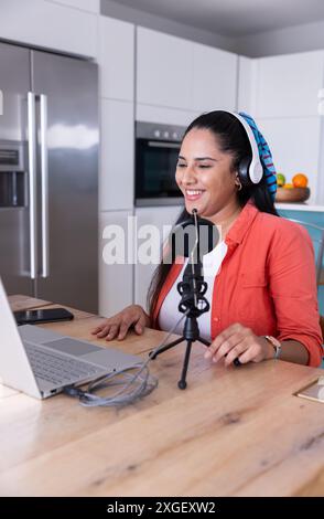 Podcasting con microfono e laptop, donna che indossa le cuffie sorridente in cucina Foto Stock