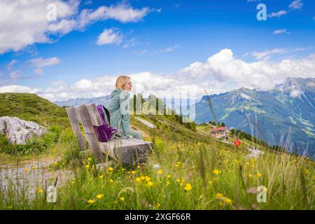 Una donna siede su una panchina in un paesaggio di montagna, circondato da un prato fiorito sotto un cielo parzialmente nuvoloso, Penken, Zillertal. Austria Foto Stock