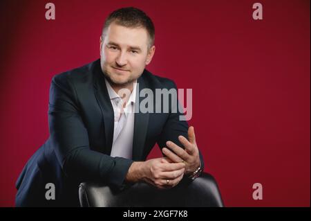 Ritratto di un attraente uomo d'affari caucasico sorridente con un vestito. Si erge nello studio su uno sfondo rosso, appoggiato su una poltrona in pelle e. Foto Stock