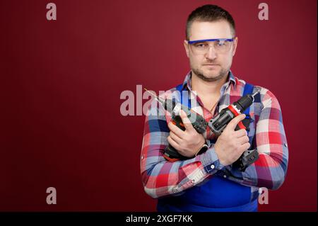 Serio riparatore caucasico in occhiali di protezione con utensili per la lavorazione del legno. Ritratto carpentiere su sfondo rosso studio. Copia spazio Foto Stock