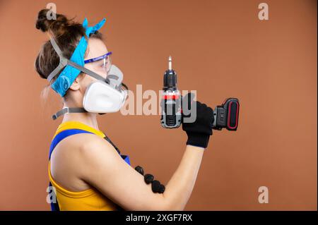 Ritratto in studio di una donna riparatrice con occhiali di protezione e respiratore con un cacciavite elettrico a batteria. Donna caucasica falegname al lavoro Foto Stock