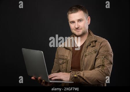 Ritratto di amichevole uomo casual caucasico positivo che utilizza un computer portatile grigio mentre si è in piedi in studio e guardando la fotocamera Foto Stock