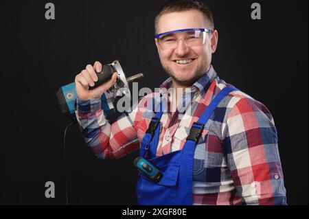 Un uomo sorridente con una tuta protettiva e una camicia a quadri che indossa occhiali di protezione per gli occhi regge e tiene in mano uno strumento di riparazione. Ritratto su nero Foto Stock