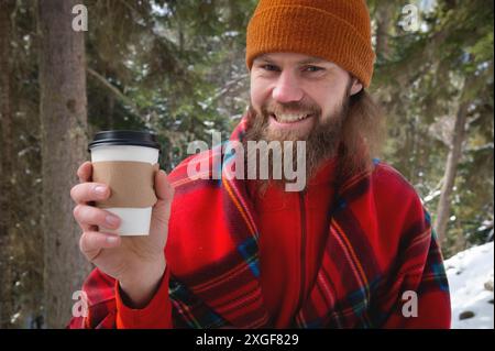 Cammina con una tazza di cioccolata calda nel parco invernale. Ritratto di un bel barbuto in abiti invernali nella neve bevendo tè o caffè caldo da un Foto Stock