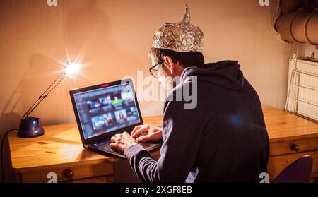 Un giovane uomo con cappuccio in alluminio è seduto nel seminterrato scuro di fronte a un computer portatile. Concetto di teoria della cospirazione Foto Stock