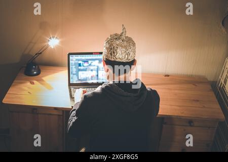 Un giovane uomo con cappuccio in alluminio è seduto nel seminterrato scuro di fronte a un computer portatile. Concetto di teoria della cospirazione Foto Stock