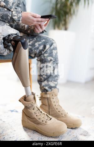 Utilizzando lo smartphone, persona in uniforme indefinita con protesi gambe sedute in casa Foto Stock
