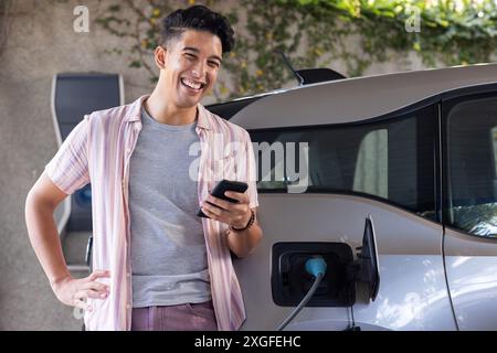 Uomo sorridente che utilizza lo smartphone mentre si ricarica l'auto elettrica dalla stazione di casa Foto Stock