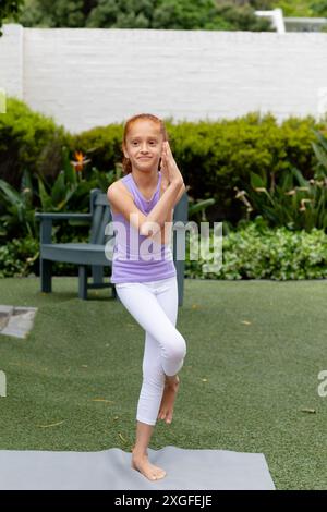 Verticale di felice ragazza birazziale in piedi su una gamba in giardino, praticando yoga, con spazio copia Foto Stock