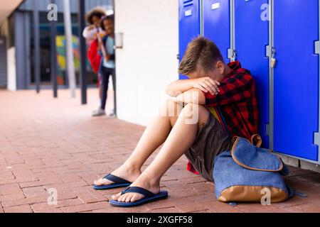 Triste scolaro caucasico seduto accanto a armadietti con scolari diversi sullo sfondo Foto Stock