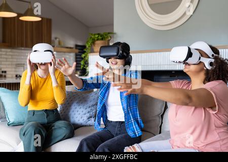 Utilizzando i visori VR, tre amiche donne sedute sul divano e che si divertono con la realtà virtuale Foto Stock