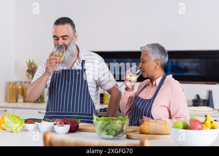 Una coppia anziana in cucina beve vino mentre prepara verdure fresche e pane Foto Stock