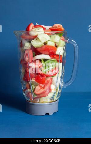 Un frullatore in vetro trasparente riempito di verdure fresche a fette, tra cui pomodori, cetrioli e peperoni su sfondo blu vivace Foto Stock