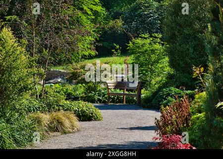Un tranquillo giardino con una panchina di legno circondata da vegetazione lussureggiante e piante varie con un sentiero che conduce ad una panchina ombreggiata da alberi, Regno Unito, Foto Stock