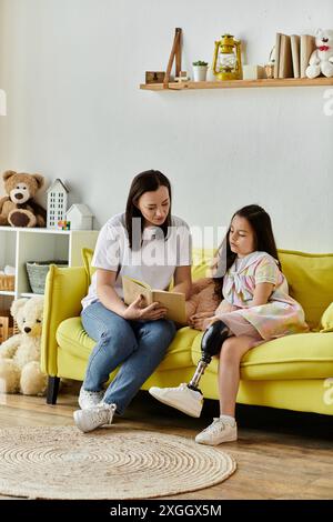 Una madre e sua figlia si siedono su un divano giallo a leggere un libro. La figlia ha una gamba protesica. Foto Stock
