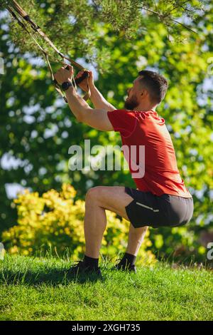 Uomo muscoloso che esegue TRX accovaglia all'aperto, utilizzando cinghie ancorate all'albero, contro una vegetazione lussureggiante. Allenamento all'aperto Foto Stock