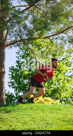 Metti in forma l'atleta che esegue esercizi TRX all'aperto con le cinghie. Uomo muscoloso che fa gli esercizi di squat nel parco verde Foto Stock