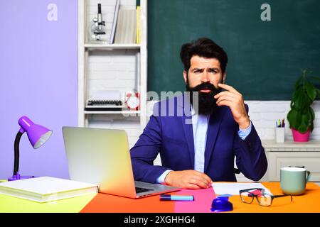 Insegnante maschile confuso o tutore universitario che si trova alla scrivania in classe. Torna a scuola. L'insegnante universitario lavora con un computer portatile. Duro lavoro. Studente in preparazione Foto Stock