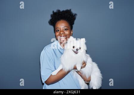 Ritratto medio di una giovane esperta di veterinaria con un cane bianco della pomerania in mano. Foto in studio di un veterinario sorridente che guarda la fotocamera isolata su sfondo blu, flash della fotocamera, spazio di copia Foto Stock