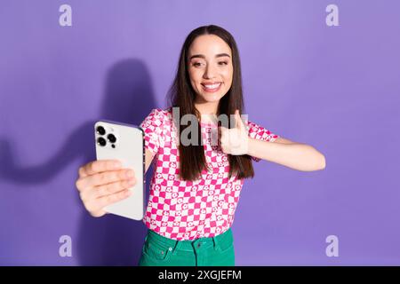 Ritratto fotografico di una giovane donna attraente che tiene il pollice verso l'alto approva abiti rosa eleganti e vestiti isolati su sfondo di colore viola Foto Stock