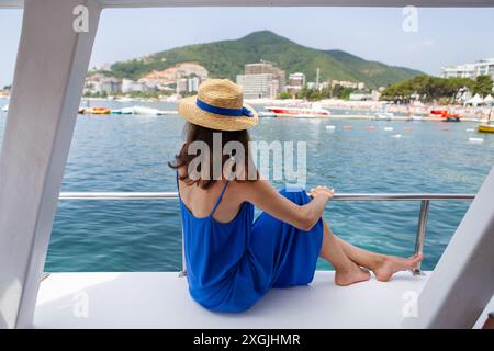 Una donna con un abito blu e un cappello di paglia si siede su una barca, osservando il paesaggio urbano costiero e le acque cristalline Foto Stock