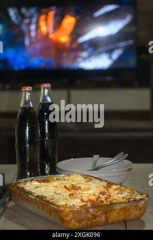 Concediti una lasagna fatta in casa servita calda e fresca dal forno, accompagnata da due bottiglie di cola. Perfetto per una serata accogliente al cinema a casa. Questa inv Foto Stock