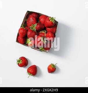 "Vista dall'alto delle fragole fresche in un piccolo contenitore di cartone, con alcune fragole sparse intorno, su uno sfondo bianco pulito. Ideale Foto Stock