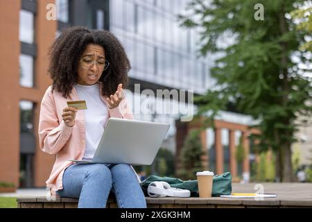 Donna con capelli ricci seduta all'aperto con carta di credito, che sembra preoccupata mentre usa il computer portatile. Concetto di shopping online, problemi finanziari e lavoro remoto. Foto Stock