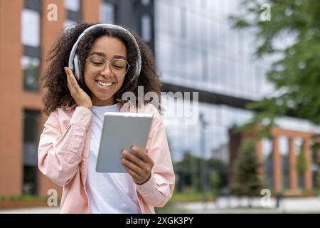 Donna sorridente con capelli ricci e occhiali che ascolta la musica con le cuffie e usa il tablet all'aperto. Si erge vicino a un edificio moderno in un'atmosfera rilassata e allegra. Foto Stock