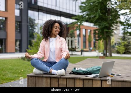 Giovane donna che meditava in uno spazio pubblico all'aperto con un portatile. Si impegna nel lavoro a distanza mentre pratica la consapevolezza. Ambiente rilassato e tranquillo con edificio moderno sullo sfondo. Foto Stock