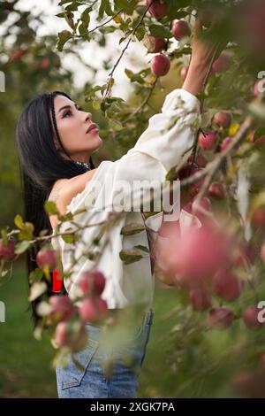 Donna concentrata e tatuata in camicetta senza spalle, che raggiunge la mela sull'albero nel frutteto. Vista laterale della giovane donna caucasica che raccoglie mele nel giardino, gli alberi sono carichi di frutti rossi. Concetto di mietitura. Foto Stock