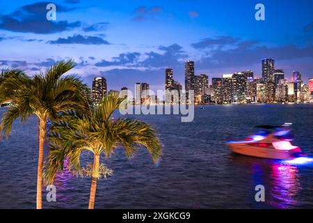 Vista notturna dello skyline di Miami in Florida attraverso Biscayne Bay con grattacieli illuminati, palme e barche Foto Stock