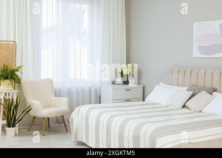 Letto grande, cassettiera, poltrona, piante d'appartamento e finestra con tende nella camera da letto. Design degli interni Foto Stock