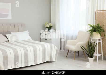 Letto grande, cassettiera, poltrona, piante d'appartamento e finestra con tende nella camera da letto. Design degli interni Foto Stock