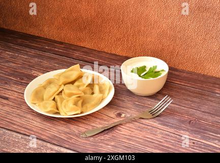 Un piatto di gnocchi appena cucinati con patate e funghi su un tavolo di legno, accanto a una forchetta e una tazza con panna acida e un rametto di prezzemolo. Cl Foto Stock