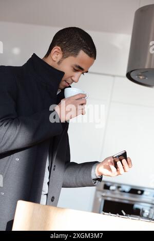 Il giovane uomo d'affari ama il caffè e controlla il telefono in una cucina moderna, bilanciando lavoro e tempo libero a casa Foto Stock