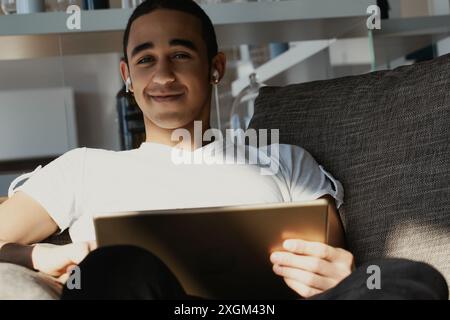 Il giovane uomo è seduto su un comodo divano nel suo salotto, si gode un po' di tempo libero con il suo tablet digitale e ascolta la musica attraverso il suo auricolare Foto Stock