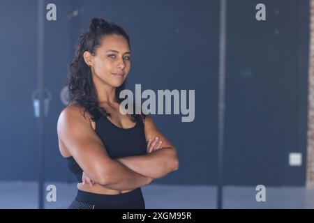 Una giovane donna birazziale forte e in forma si trova in palestra con fiducia, trasudando forza e determinazione in un ambiente fitness inalterato. Foto Stock