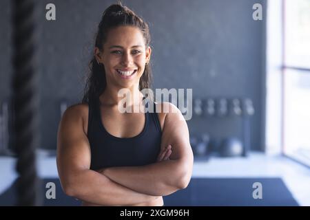 Una giovane donna birazziale forte e in forma posa con fiducia in palestra. La sua costruzione e il suo sorriso suggeriscono una dedizione al fitness e alla salute inalterata. Foto Stock