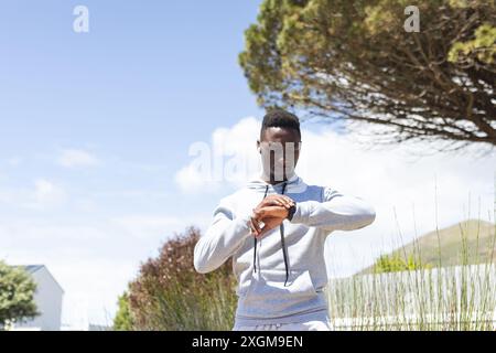 Felice uomo afroamericano che indossa felpa con cappuccio e auricolari che corrono e controllano lo smartwatch. Jogging, fitness, esercizio fisico, tecnologia e stile di vita sano, Foto Stock