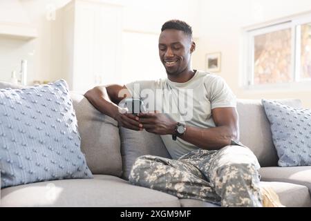Felice soldato afroamericano seduto sul divano a casa con lo smartphone e sorridente. Comunicazione, servizio militare, esercito, ritorno, casa, domicilio Foto Stock