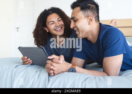 La coppia birazziale gay gode di un tablet a letto a casa. Condividono un momento di svago e connettività digitale, accogliente nella loro camera da letto. Foto Stock