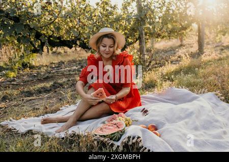 Una donna con un vestito rosso siede su una coperta in un campo, tenendo una fetta di anguria Foto Stock