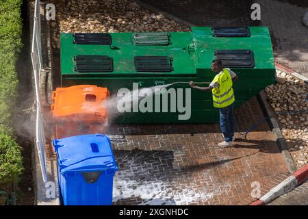 Eilat, Israele - 9 giugno 2024: Lavoratore addetto alla pulizia delle strade in strada con lavavetri riflettenti con lavavetri ad alta pressione. Contenitori per rifiuti per la raccolta differenziata Foto Stock