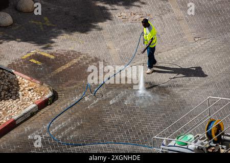 Eilat, Israele - 9 giugno 2024: Lavoratore addetto alla pulizia delle strade in strada con lavavetri riflettenti con lavavetri ad alta pressione. Contenitori per rifiuti per la raccolta differenziata Foto Stock