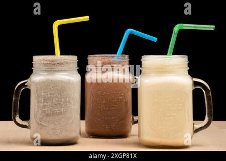 I tre bicchieri mason jar riempiti di frullati e cannucce diversi su un tavolo in legno su sfondo nero Foto Stock