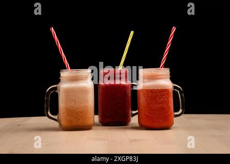 I tre bicchieri mason jar riempiti di frullati e cannucce diversi su un tavolo in legno su sfondo nero Foto Stock
