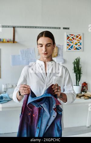 Un giovane uomo con una camicia bianca esamina attentamente un capo in denim nel suo studio di restauro dell'abbigliamento fai da te, concentrandosi sulla sostenibilità dando nuova vita Foto Stock