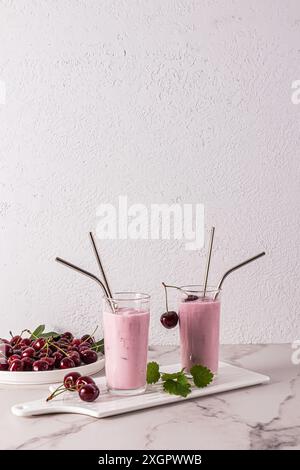 Due bicchieri alti di vetro con frullato di ciliegia su un vassoio di ceramica e una ciotola piena di ciliegie. tavolo in marmo. Delizioso dessert. Vista verticale anteriore. Foto Stock