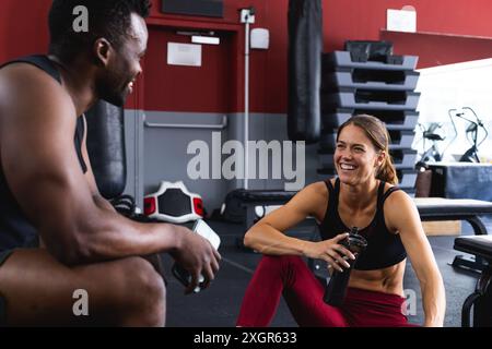 Fit uomo afroamericano e giovane donna caucasica condividono una risata in palestra. La loro interazione amichevole mette in evidenza l'aspetto sociale dell'ambiente fitness Foto Stock